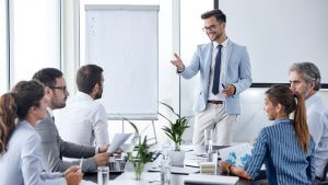 People sitting in an office meeting at a table with the CEO standing in the front leading the meeting