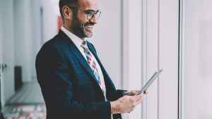 Cheerful prosperous businessman 50 years old dressed in formal wear holding digital tablet to check mail while looking out of window