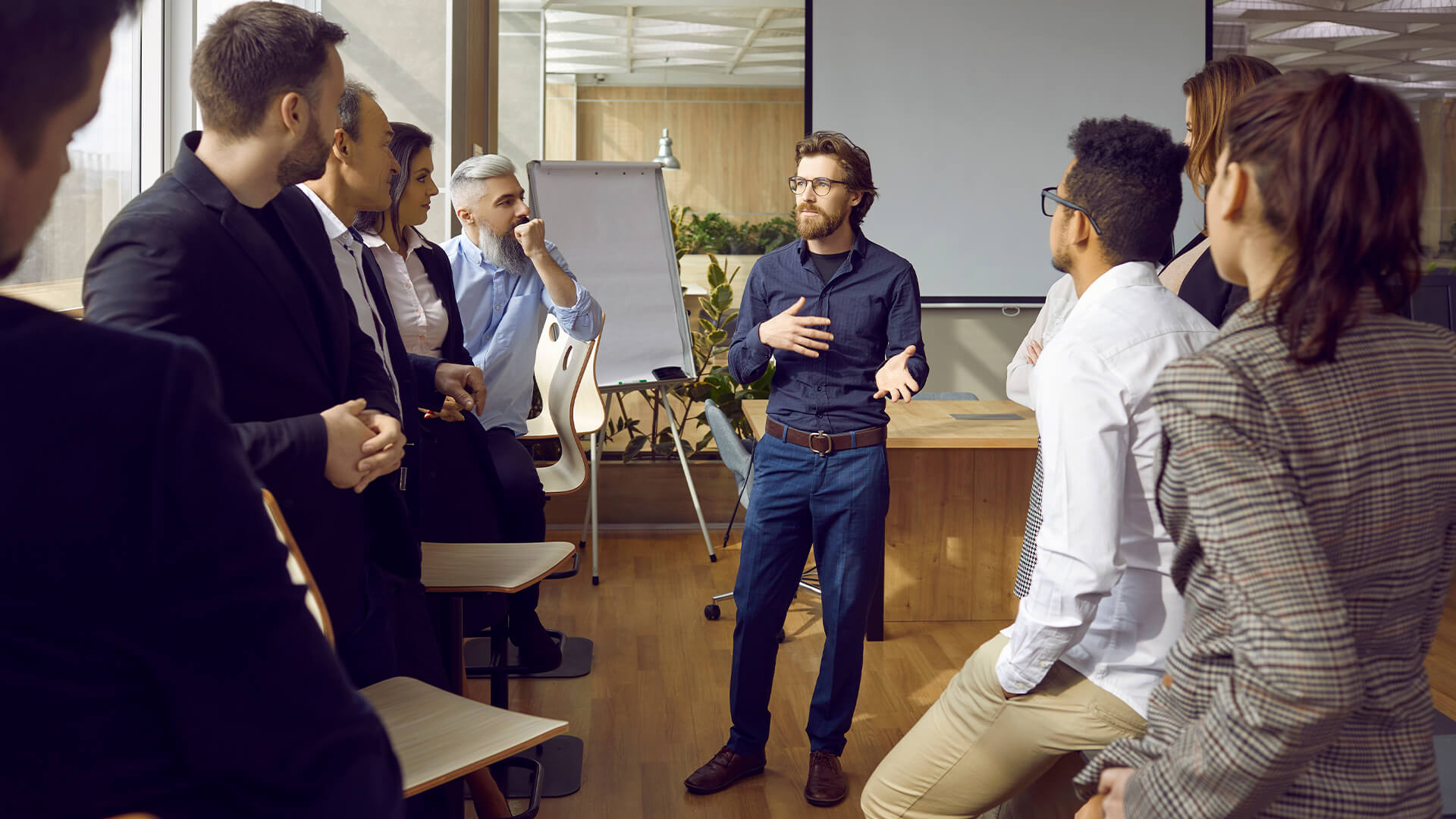 Serious young man business trainer talking to group of people in office standing around him and listening.