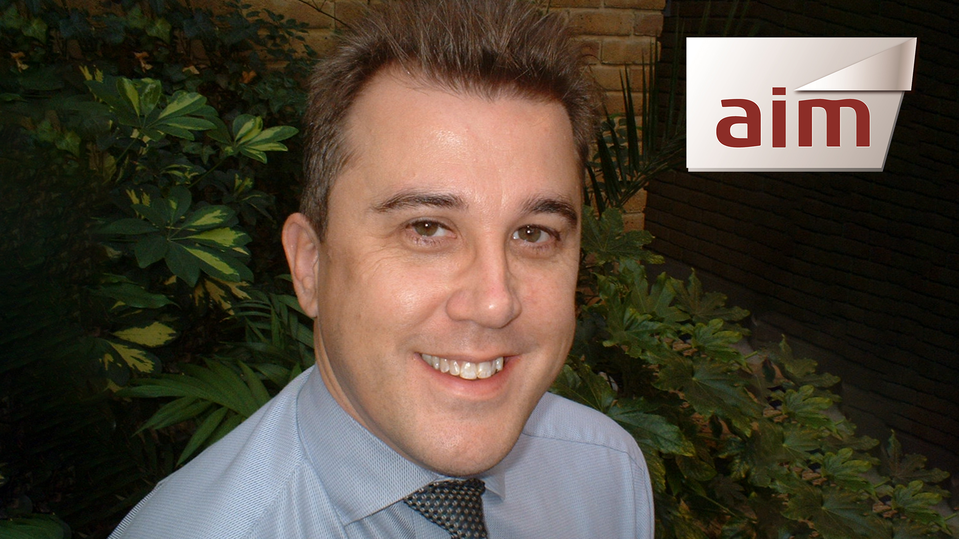 man in shirt and tie smiling for photo in front of brick wall and plants